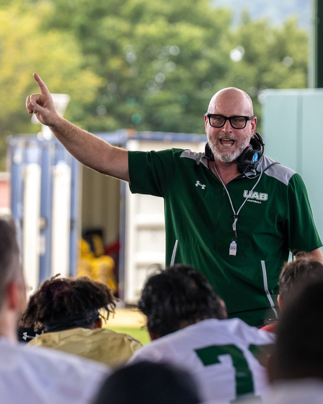 Coach or trainer giving instructions during a football practice or game, wearing a green shirt with 'UAB Football' logo, headset around neck, pointing and speaking to players.