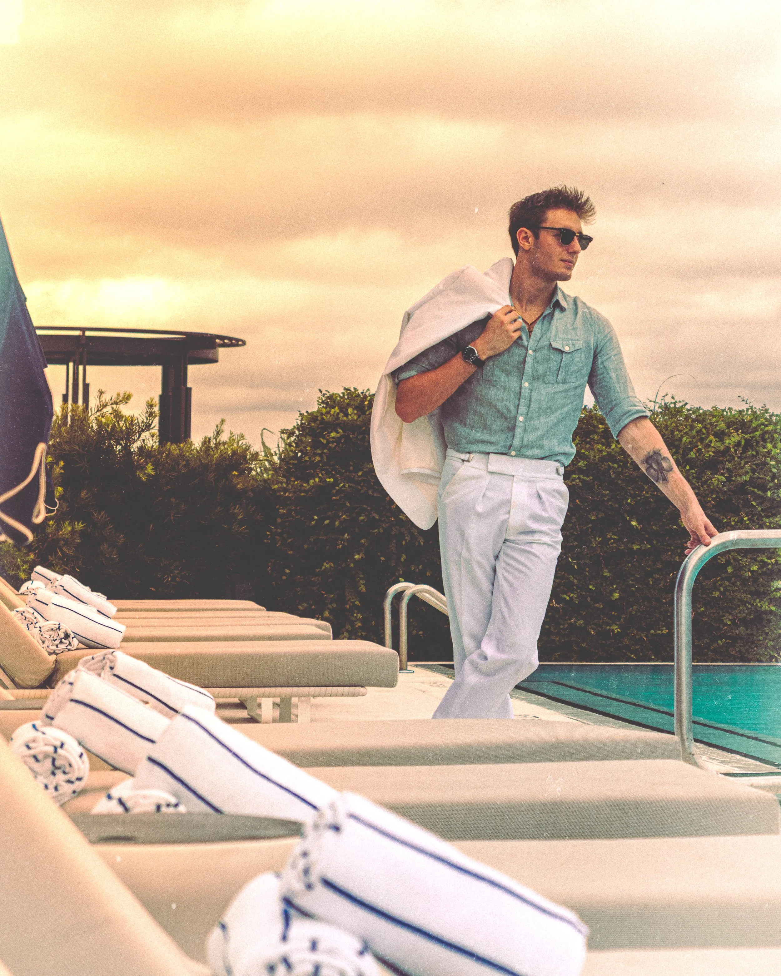 A young man in sunglasses and light-colored clothing walking by a poolside with lounge chairs and rolled towels.