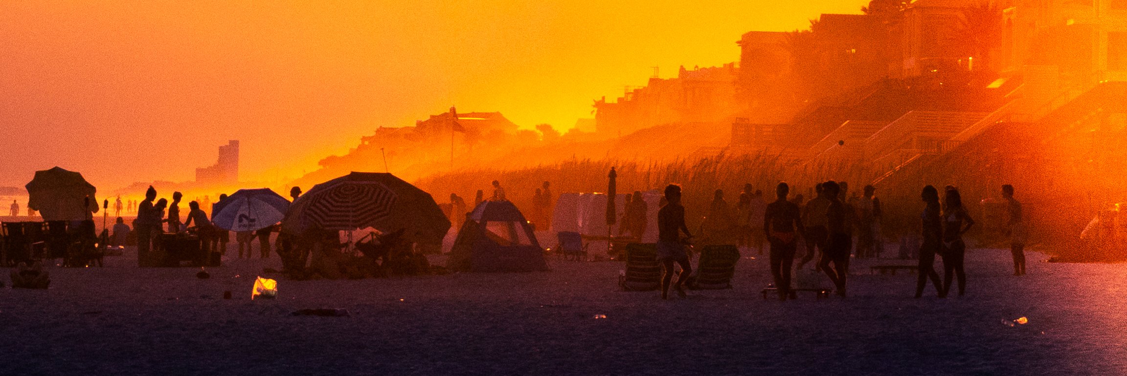 People gathered on a beach at sunset, silhouettes visible against an orange sky, some under umbrellas, with tents and buildings along the shoreline.