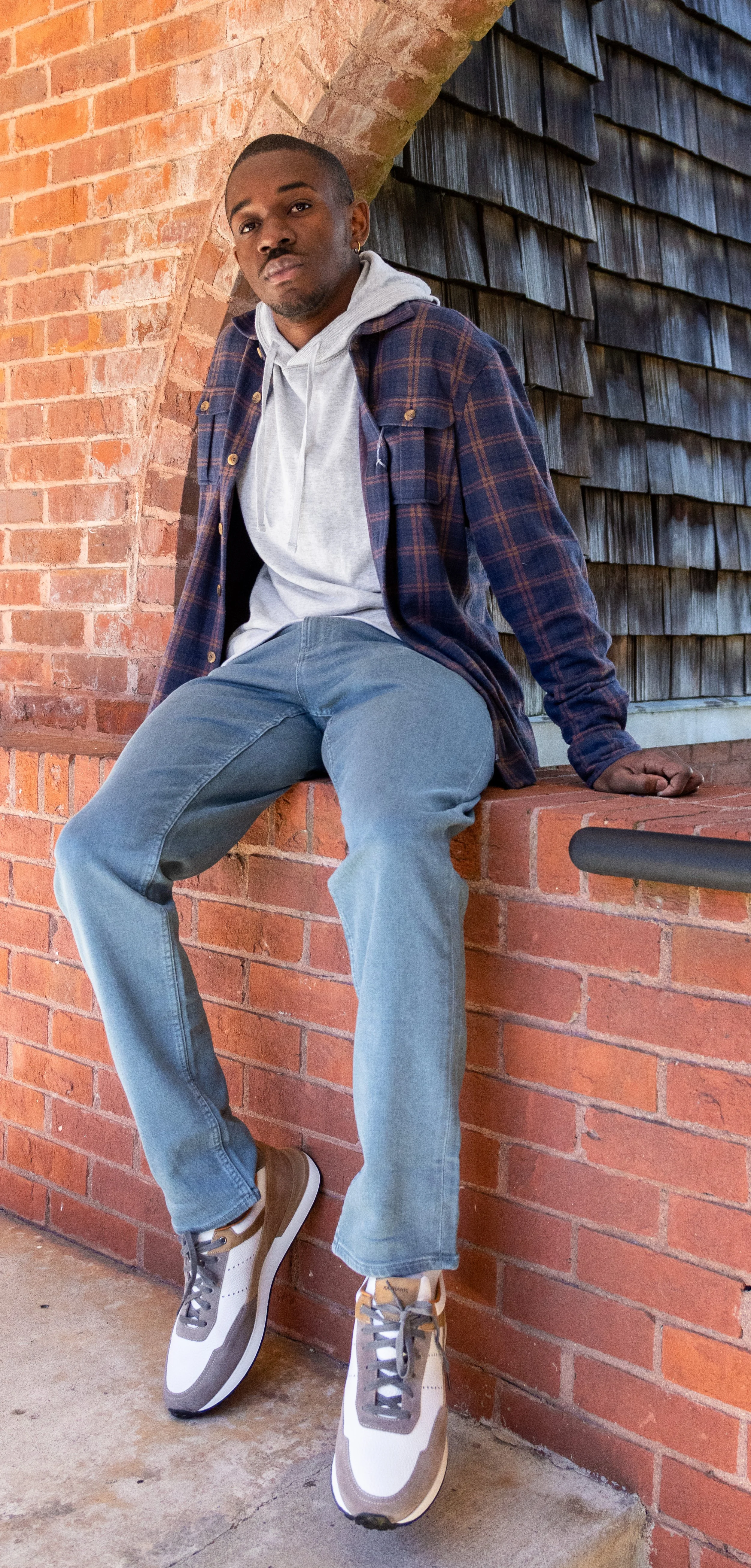 A young man with dark skin and short hair, wearing a gray hoodie under a blue and red plaid shirt, blue jeans, and sneakers, sitting on a brick ledge against a brick and wood wall background.