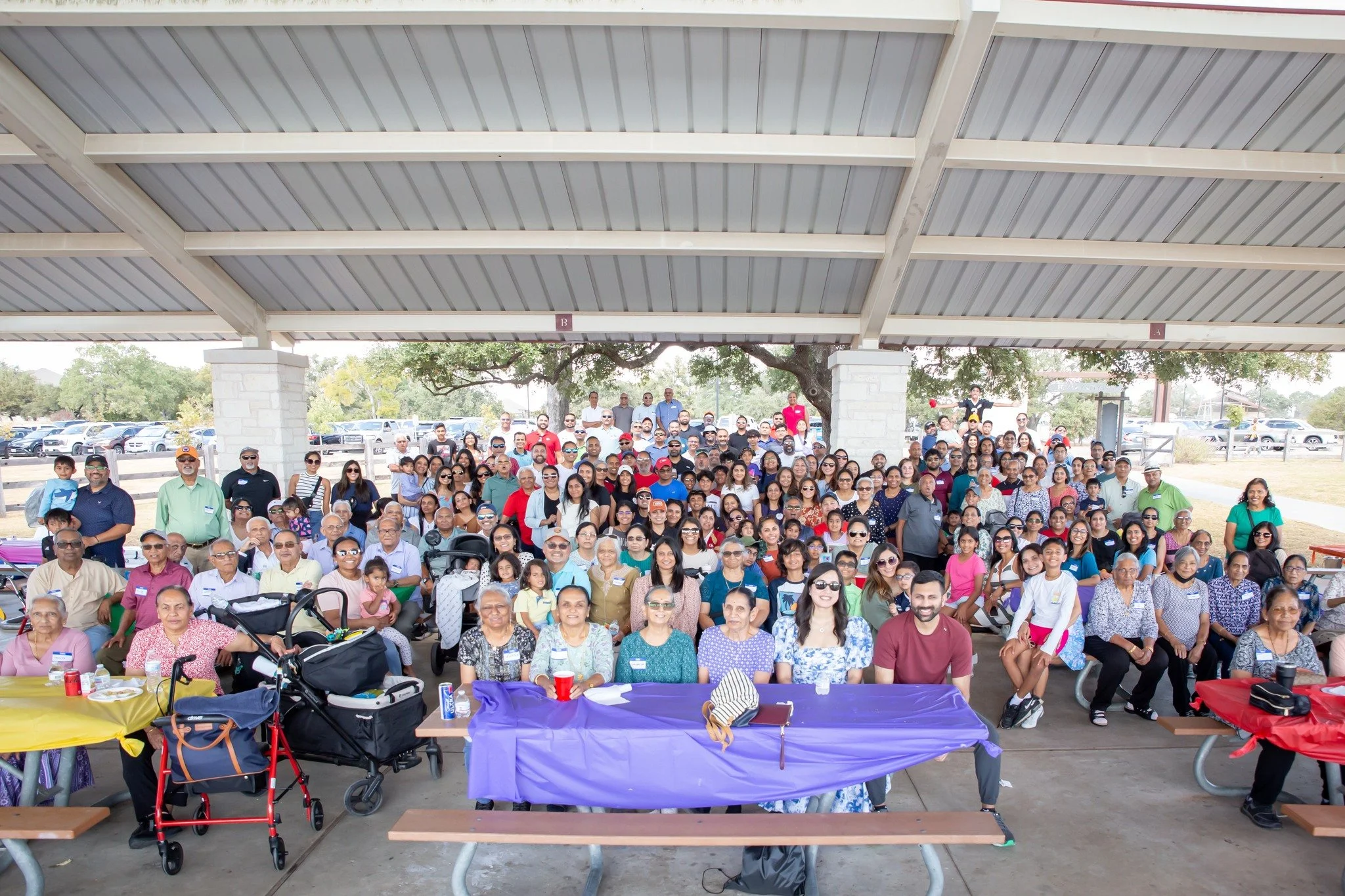 Group of diverse people gathered under a large pavilion, sitting and standing for a group photo, with trees and parked cars in the background.