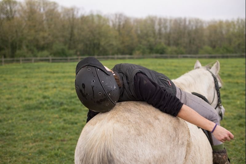 Person wearing a black helmet lying on a white horse in a grassy field.