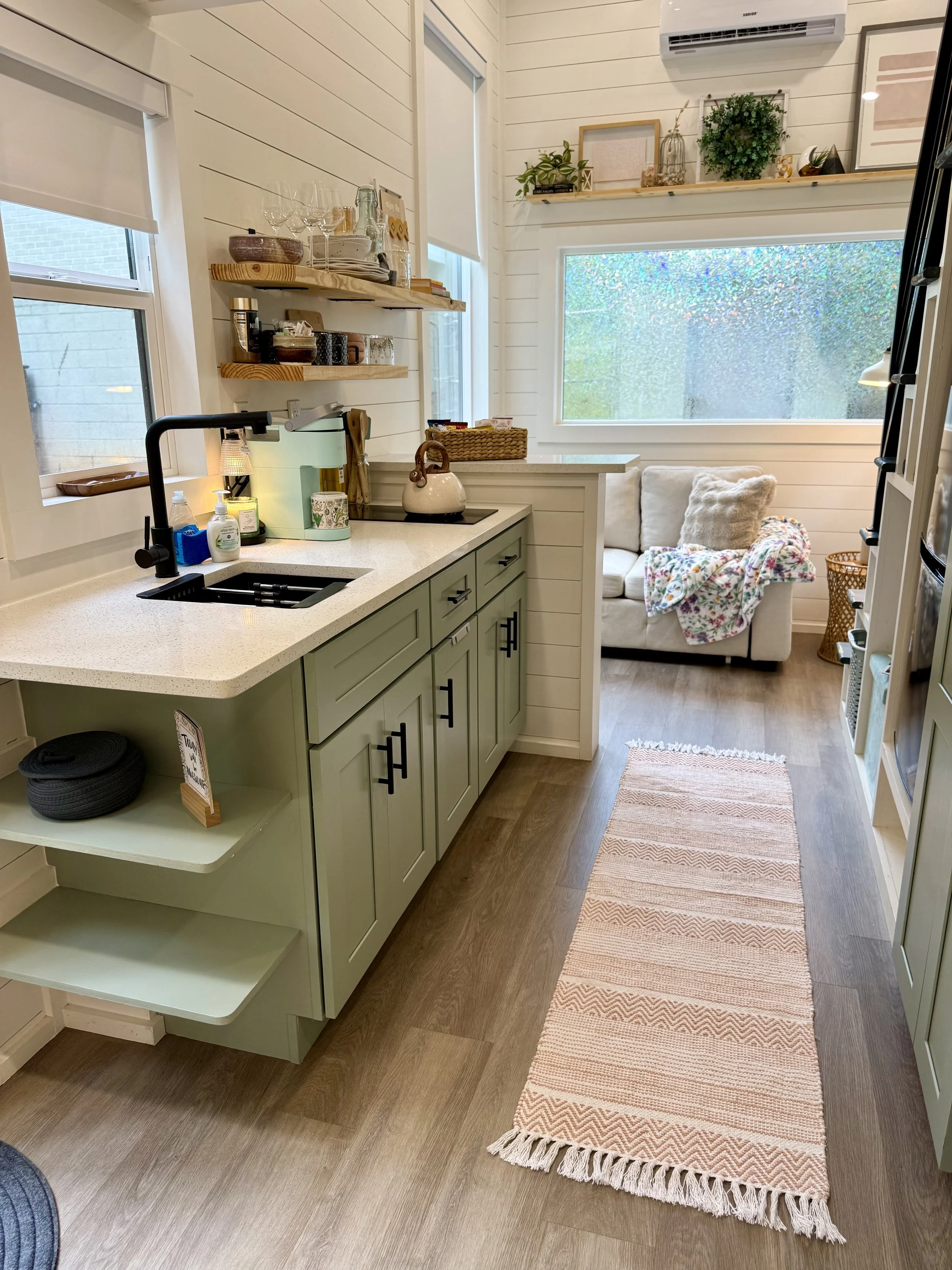 Kitchen with green cabinets, a white countertop with a black sink and faucet, open shelves with dishes and glasses, a small countertop appliances, and a living area with a beige couch, a blanket, and a window with a view of greenery outside.
