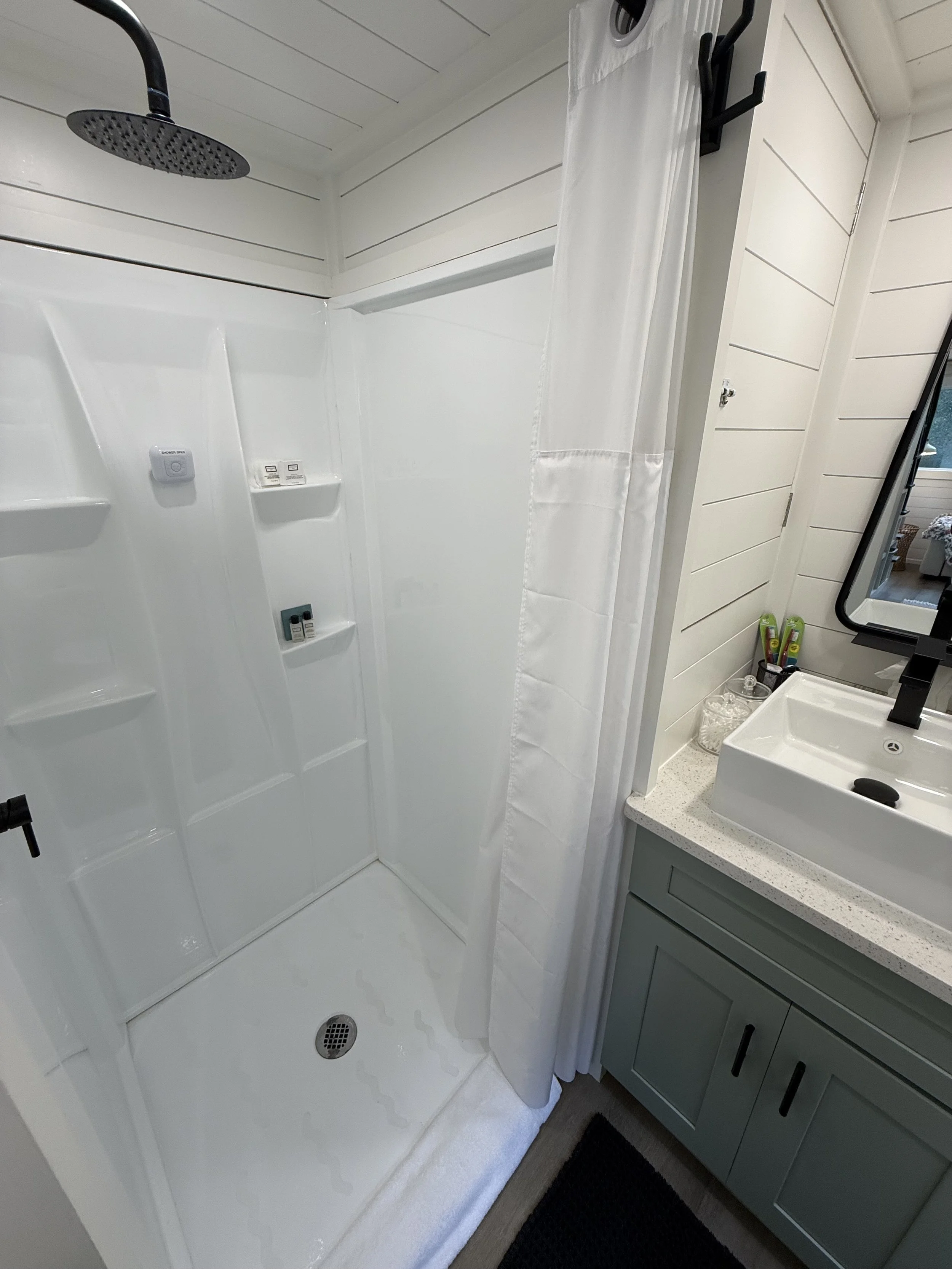 A compact bathroom with a white shower area featuring a wall-mounted showerhead, shelf with toiletries, a white curtain, and a vanity with a sink, mirror, and various toiletries on the countertop.