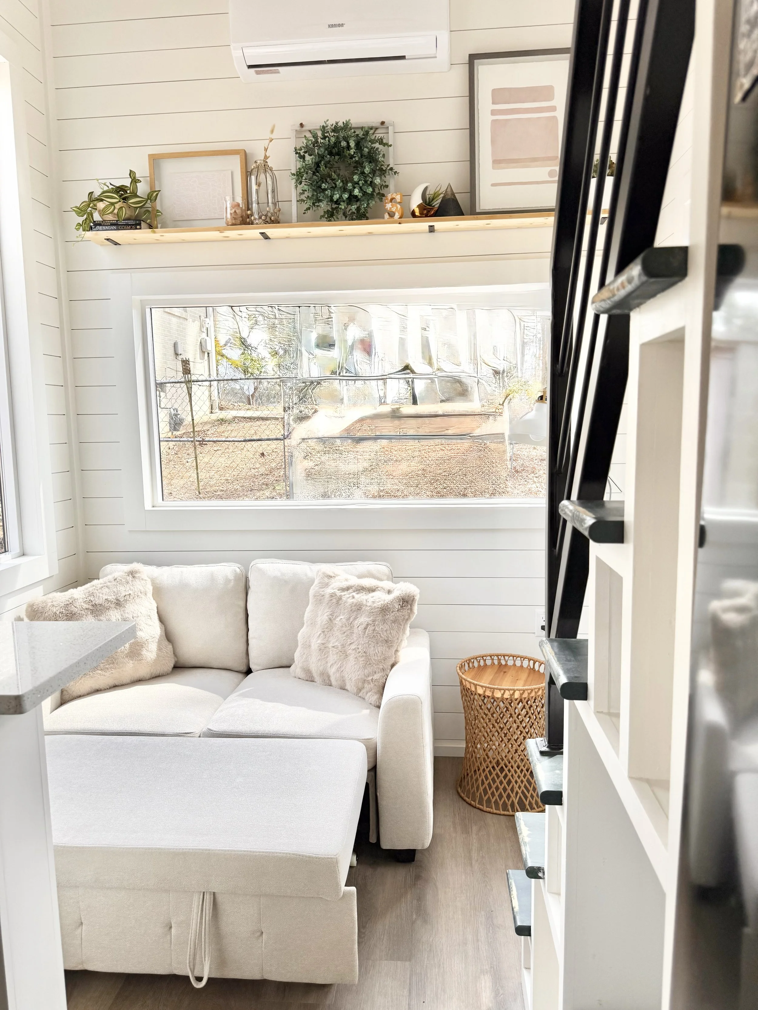 A cozy living room corner with a white sofa having plush beige pillows, a woven basket beside it, a window with a view of the outdoors, a wall-mounted air conditioner, and a decorative shelf with plants and framed artwork.