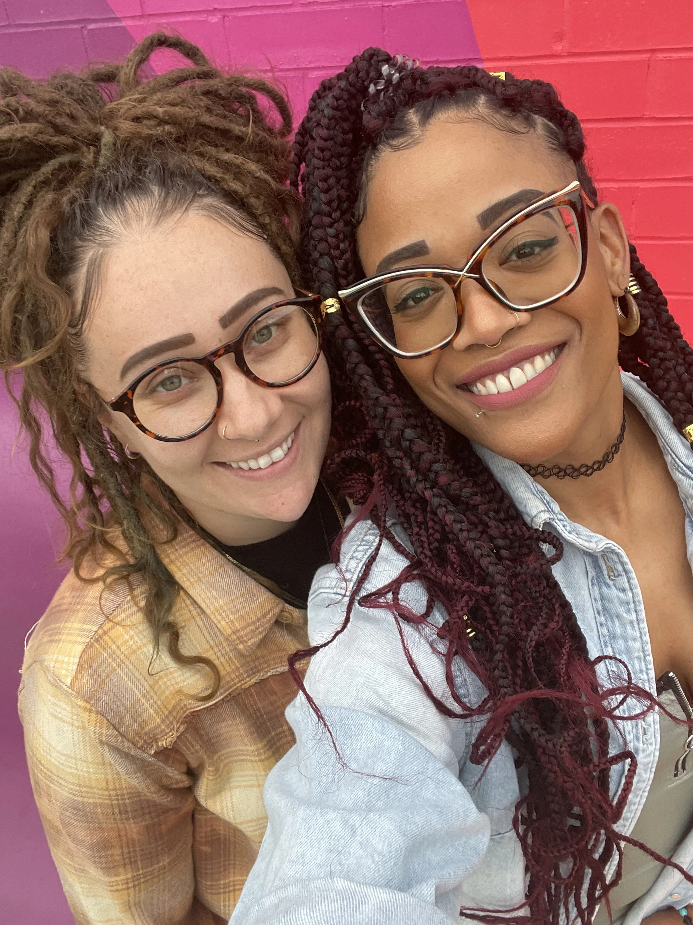 Two smiling women wearing glasses, one with dreadlocks and one with braids, close together in front of a pink wall.