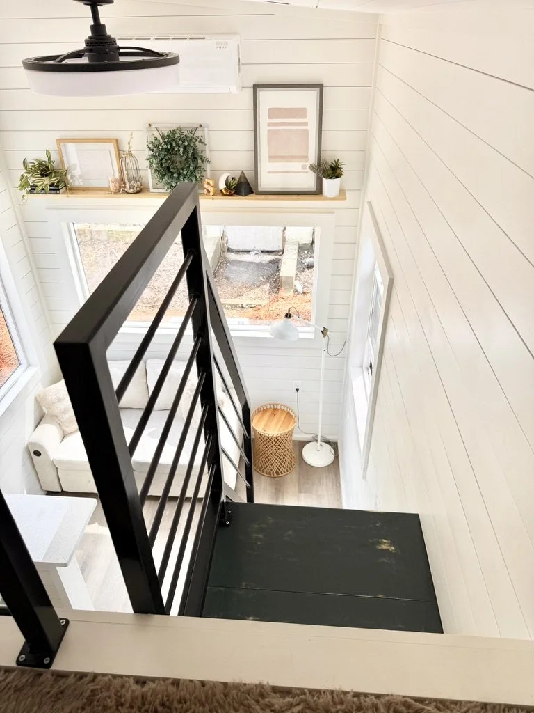 View from a rustic staircase overlooking a bright living room with white shiplap walls, a large window, a beige sofa, a white floor lamp, decorative framed art, and a shelf with plants and decor. Outside the window, a yard space is visible.