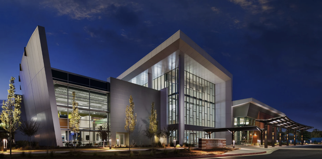 Modern building with glass windows and angular design at dusk, with trees and exterior lighting.