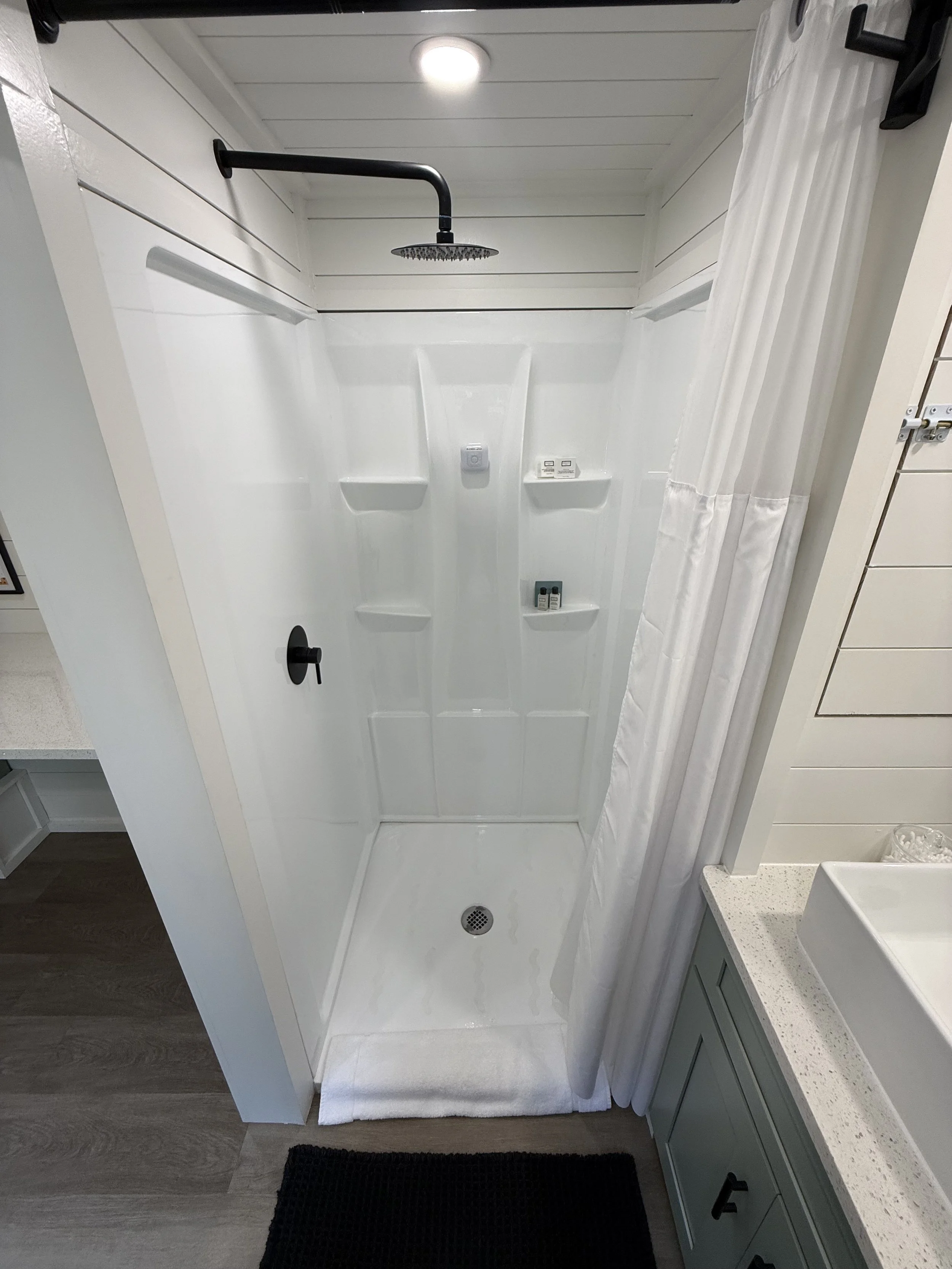 A modern shower area with a black showerhead and handle, a white shower curtain, built-in shelves holding toiletries, and a white floor mat. To the right, there is a sink with a white countertop and green cabinets.