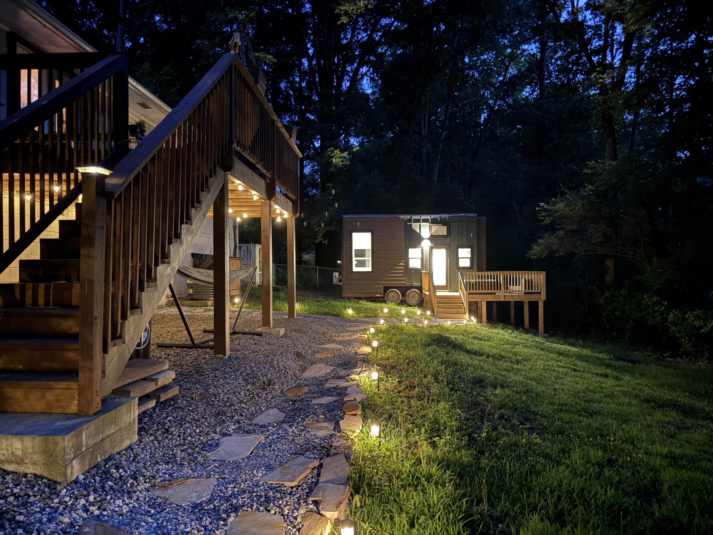 Nighttime backyard view showing a wooden staircase leading to a deck, with string lights underneath, a small modern trailer house with lights on, and a wooded area with trees in the background.