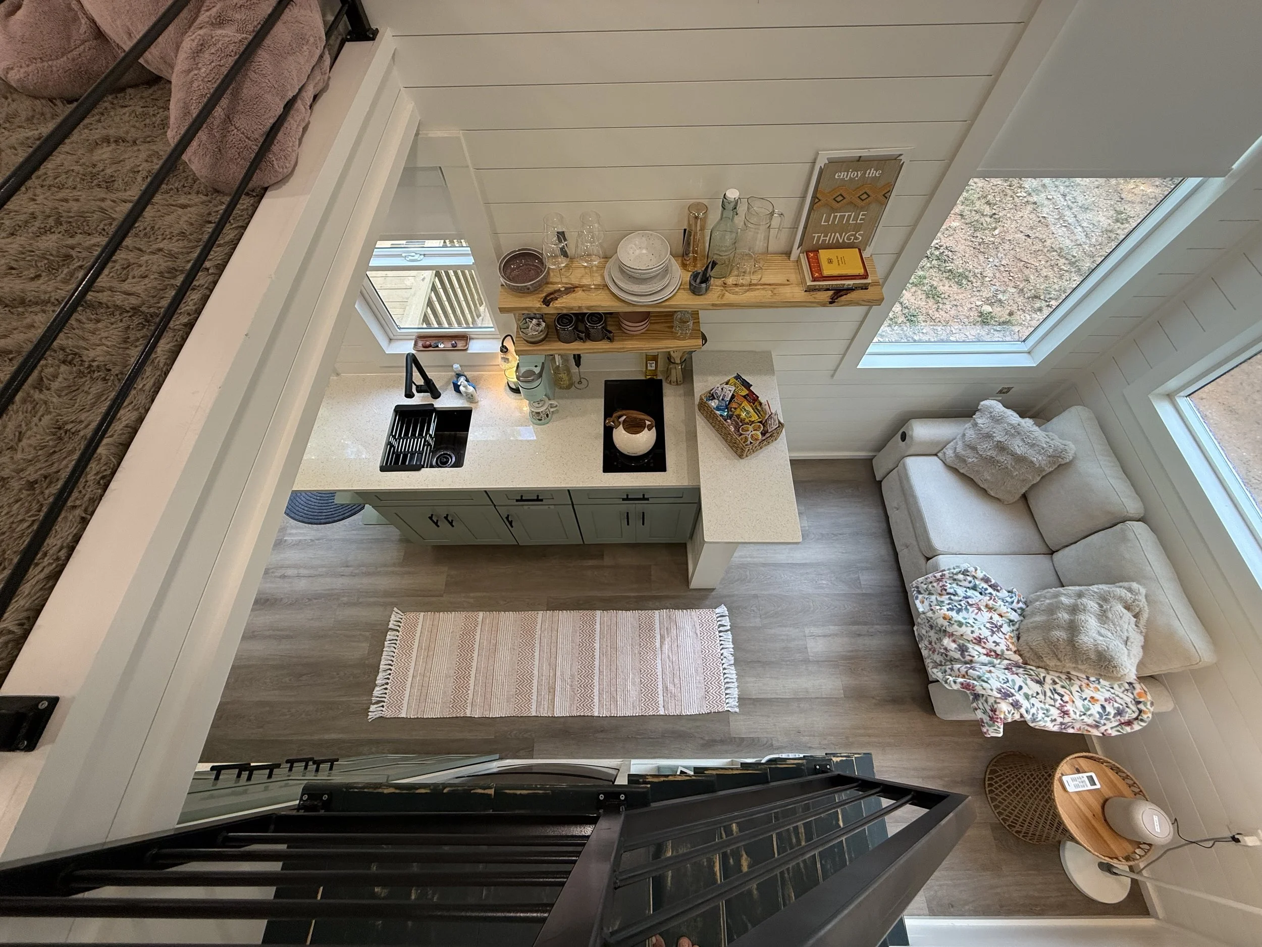 View of a cozy living space from stairs, featuring a kitchen with a white countertop, a sink, a small window, a shelf with dishes, glasses, and books, a framed sign that says 'enjoy the little things,' a beige armchair with a floral pillow, a small side table with a lamp and a fan, and large windows showing an outdoor view of dirt ground and trees.