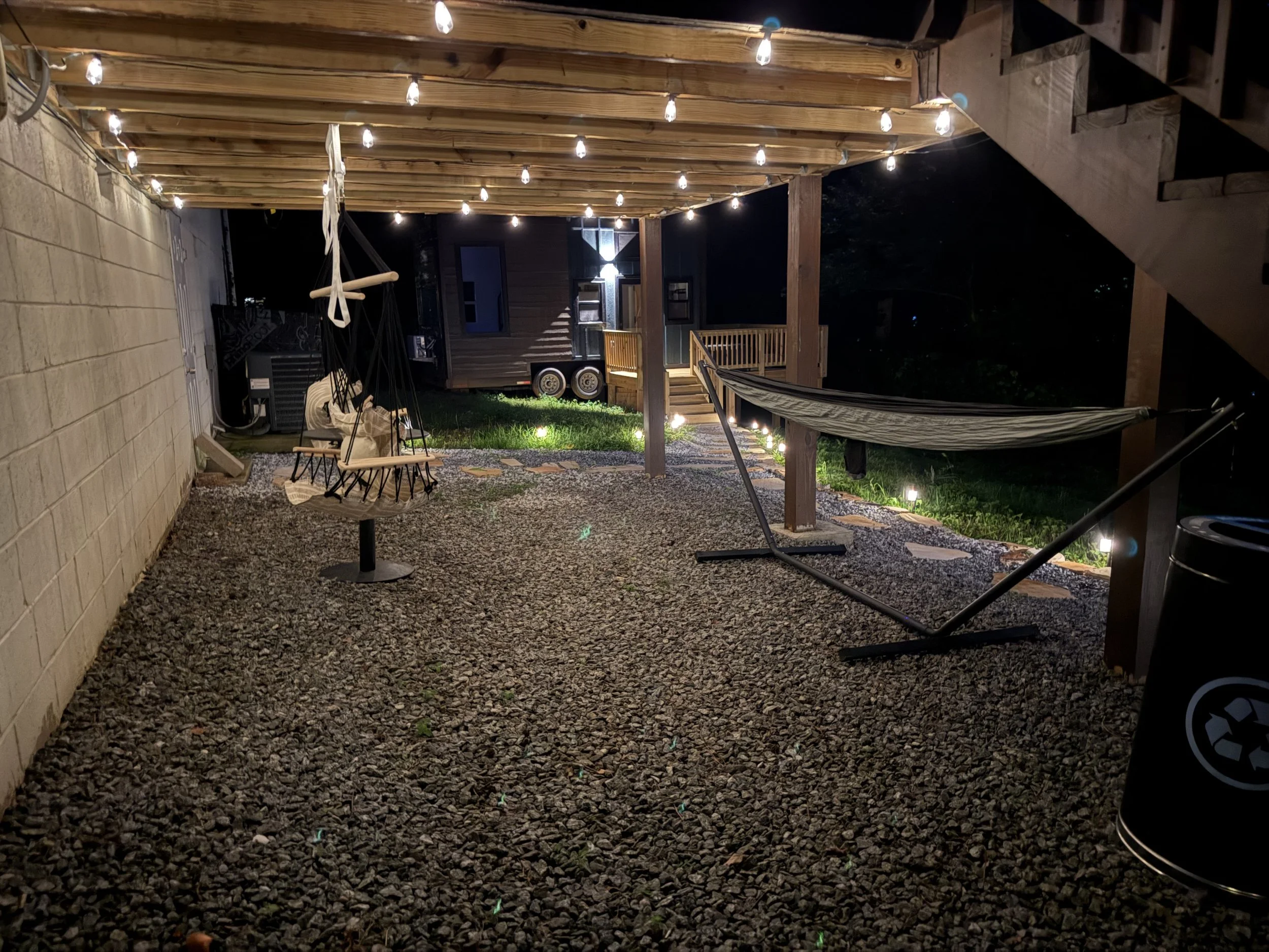 Backyard patio at night with string lights hanging from wooden beams, a swing and hammock, small grassy area, gravel ground, and a small mobile home or trailer in the background.