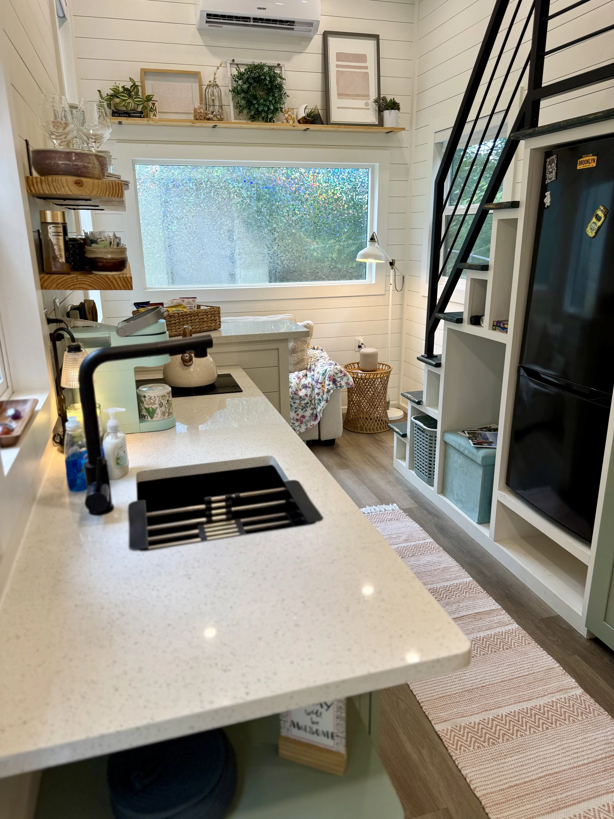 A cozy kitchen with a white countertop, black sink, and soap dispensers. Behind the counter is a white bookshelf, a fridge, a staircase, and a window with textured glass. Decor includes framed pictures, plants, and a small lamp.
