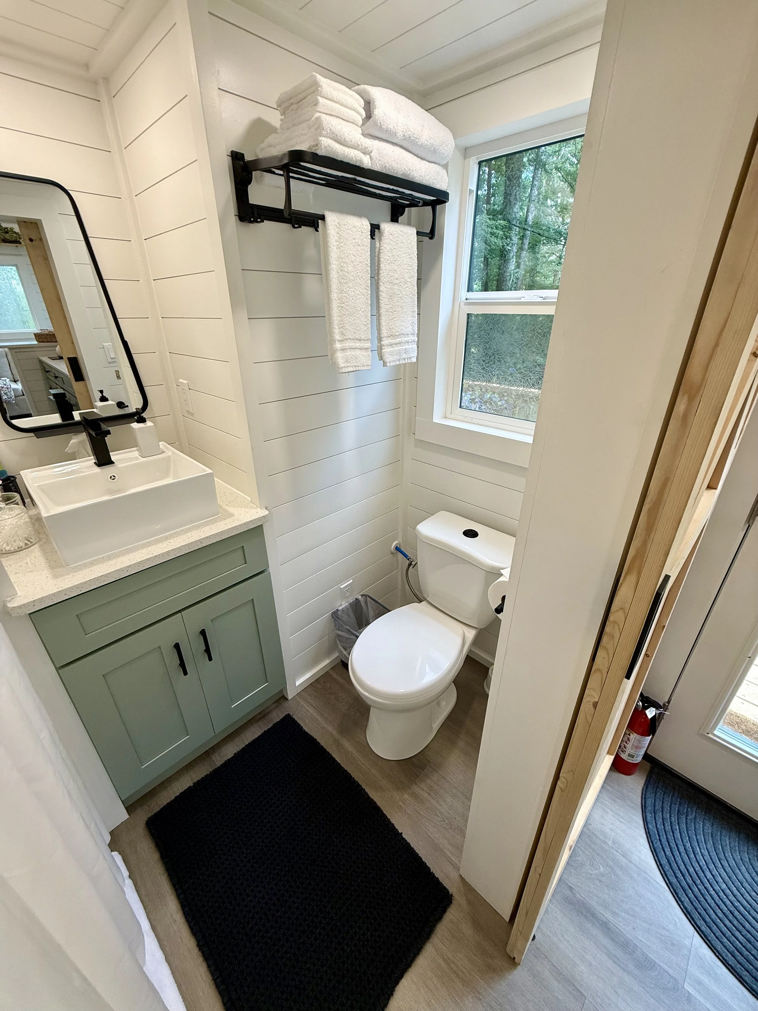 A small bathroom with a green vanity sink, a white countertop, a black framed mirror, a white toilet, a window with greenery outside, a black towel rack with white towels, a black rug on light wood flooring, and a corner with fire extinguisher and wooden beams.