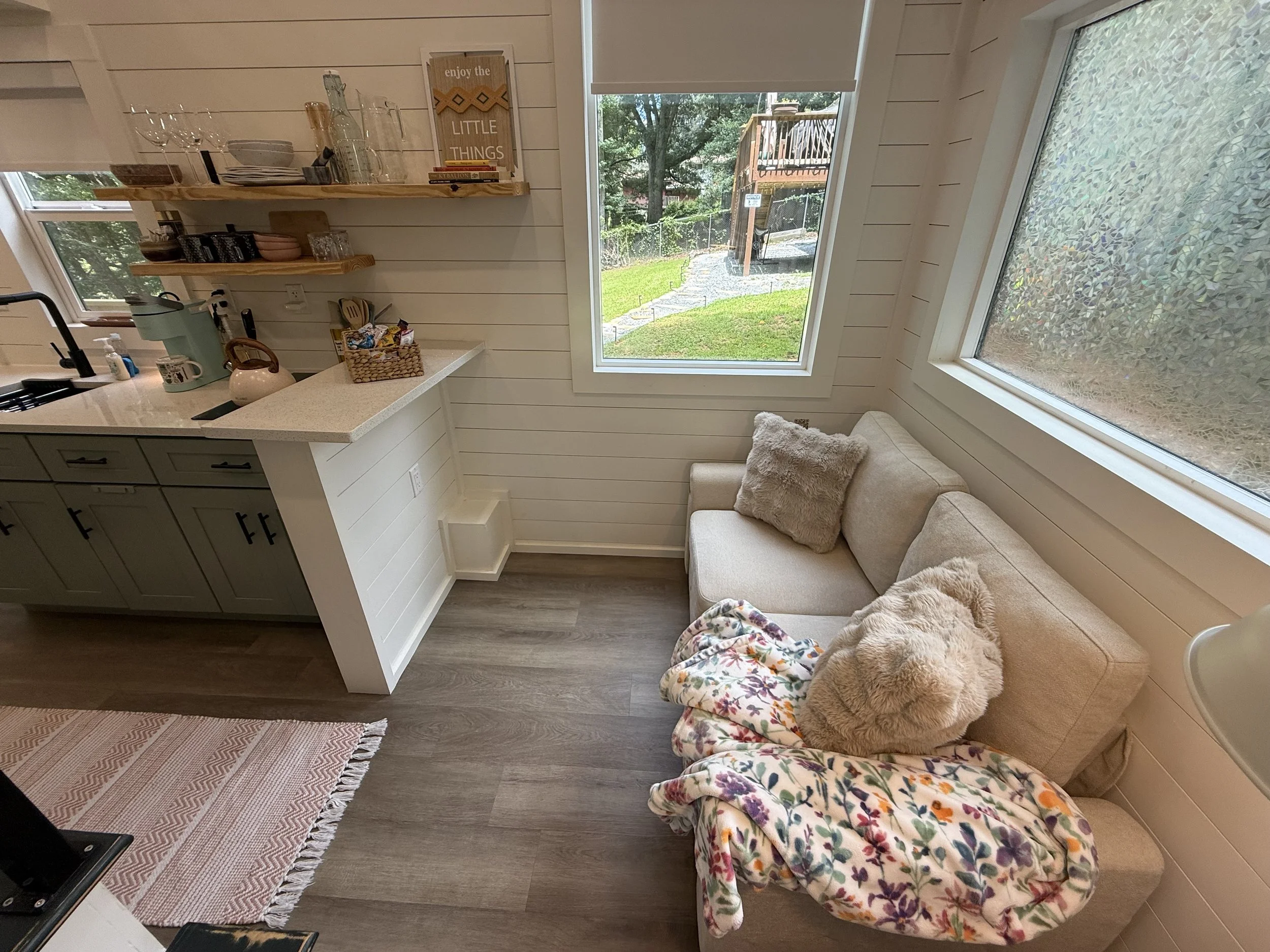 Interior of a cozy kitchen nook with a beige couch draped with a floral blanket and pillows, large windows showing a backyard with a deck, tree, and lawn, open shelving with dishes and a framed quote, and a kitchen counter with a basket of snacks and a kettle.