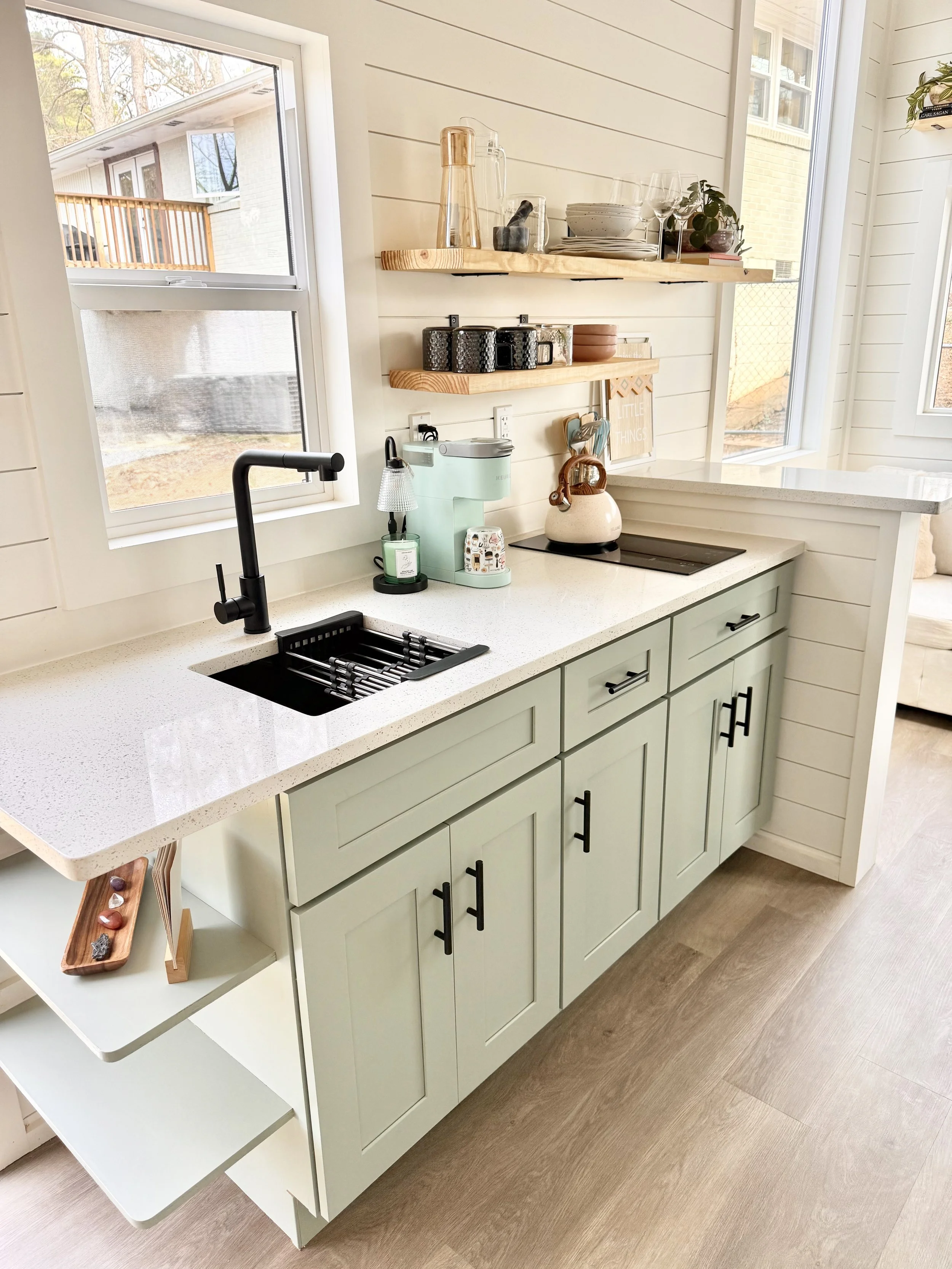 A kitchen with pale green cabinets, a white countertop with a built-in black sink and black faucet, and open shelving above. The shelves hold plates, bowls, glasses, and decorative items. There is a window above the sink and another to the right, let
