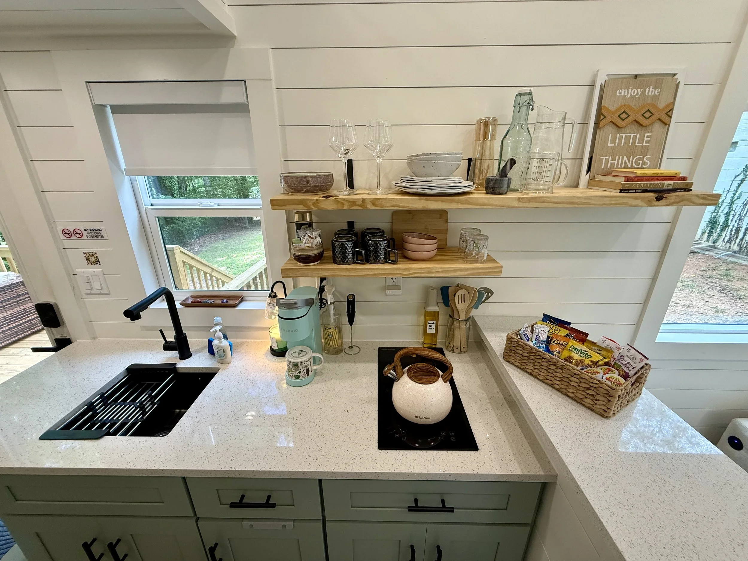 A kitchen countertop with a black sink, a white kettle, and various small kitchen items. Above the counter are two wooden shelves holding glassware, bowls, plates, a framed sign that says 'enjoy the LITTLE THINGS,' and books. A window shows a backyard with trees, and a sliding glass door is to the right.