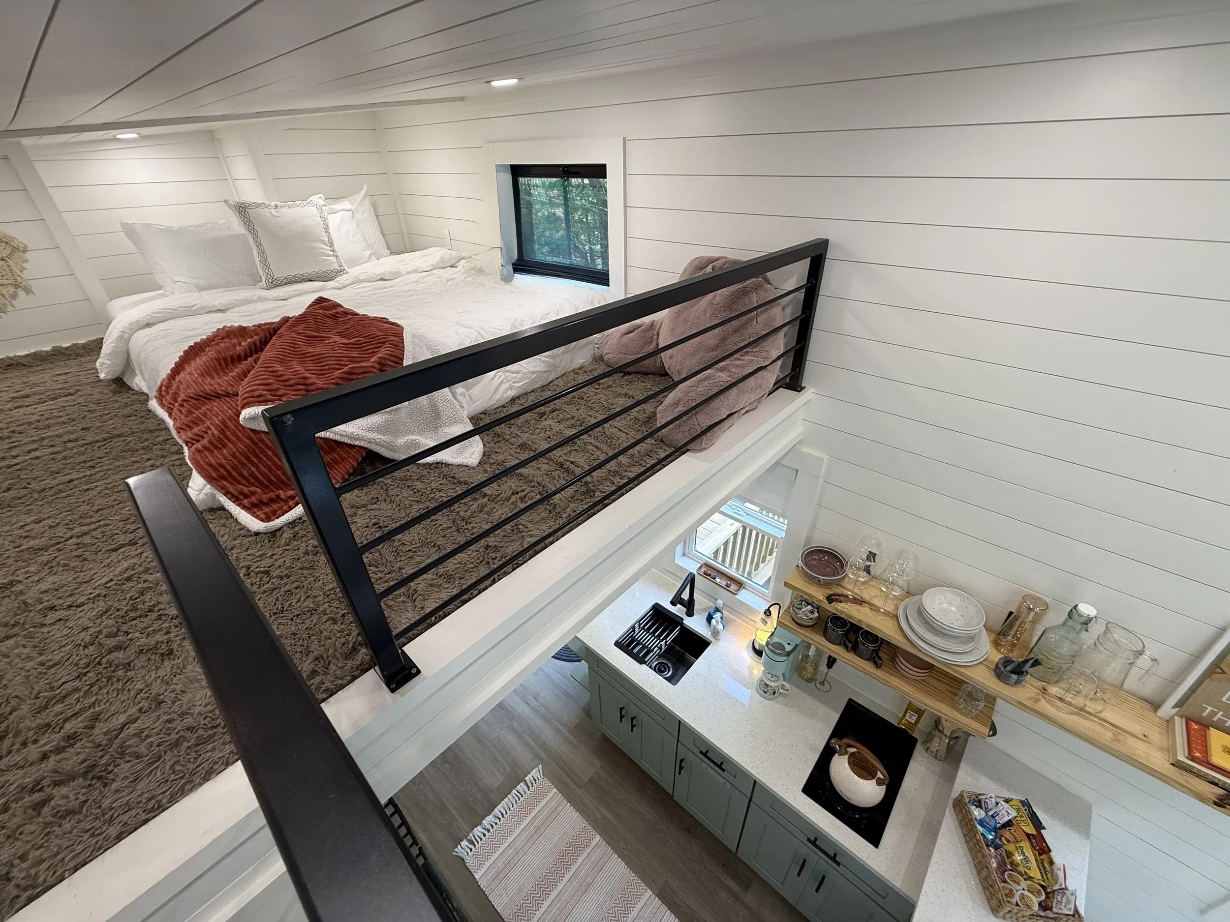 A cozy bedroom with white shiplap walls and ceiling, a bed with white bed linens and pillows, a rust-colored throw blanket, a brown shaggy rug, and a small window with greenery outside. There is a black metal headboard and a nearby kitchen area with light green cabinets, a white countertop, and open shelving holding dishes and glassware.