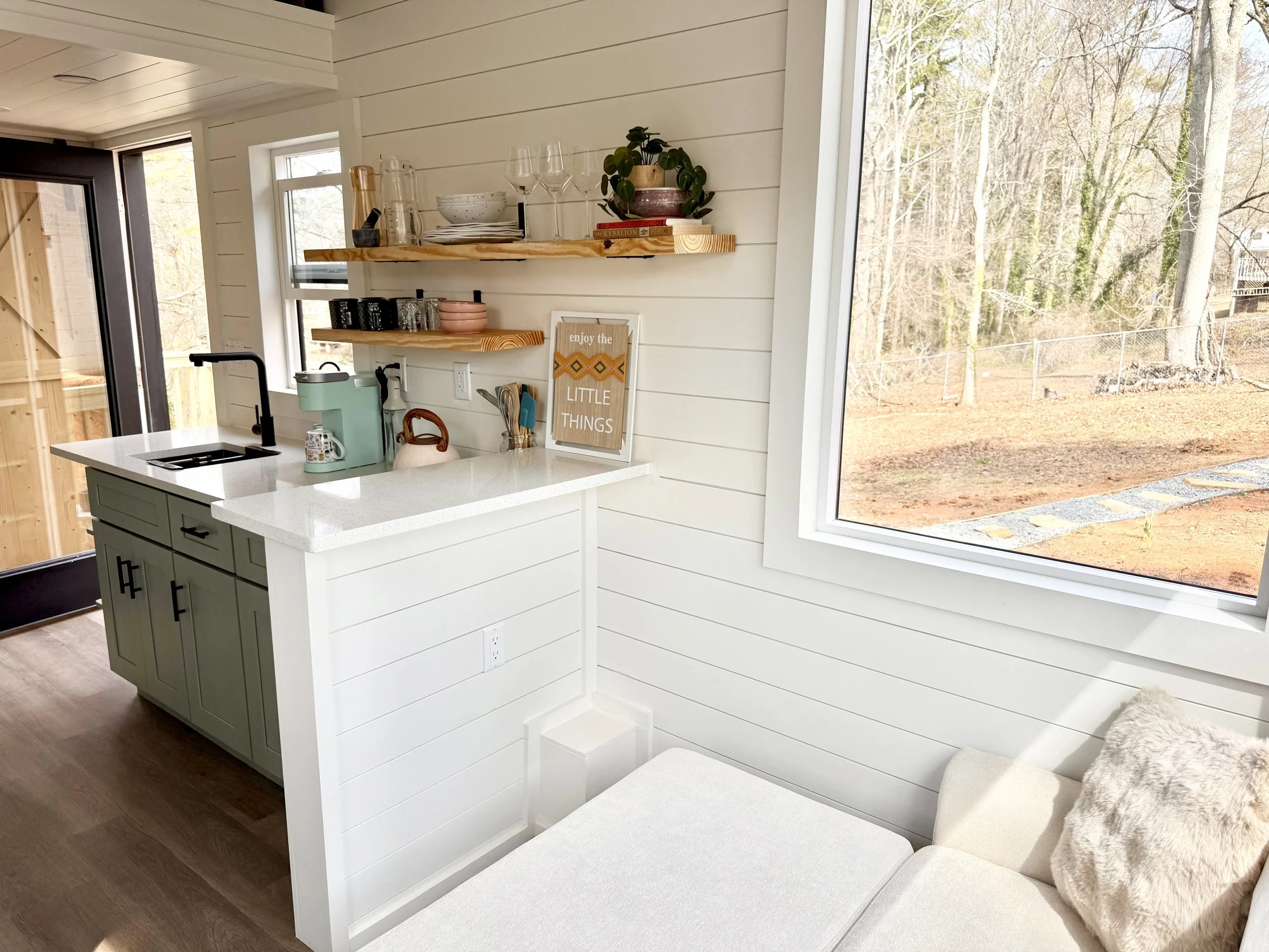 A cozy kitchen corner with a white countertop, green cabinets, floating wooden shelves with glassware and decor, a window showing outdoor trees and yard, and a small seating area with a beige cushion and fluffy pillow.