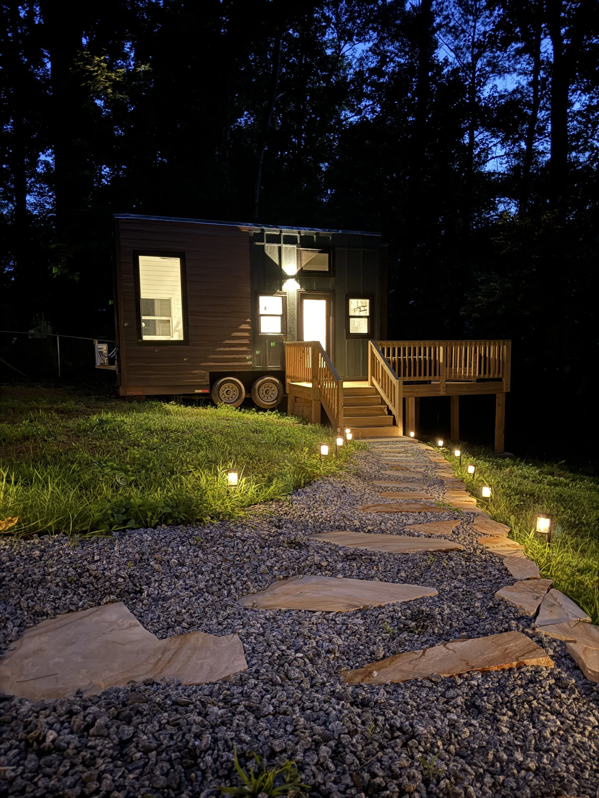 A small, dark-colored tiny house on wheels illuminated by exterior lights, with a wooden deck and a pathway with small lights leading to it, surrounded by green grass and trees at dusk.