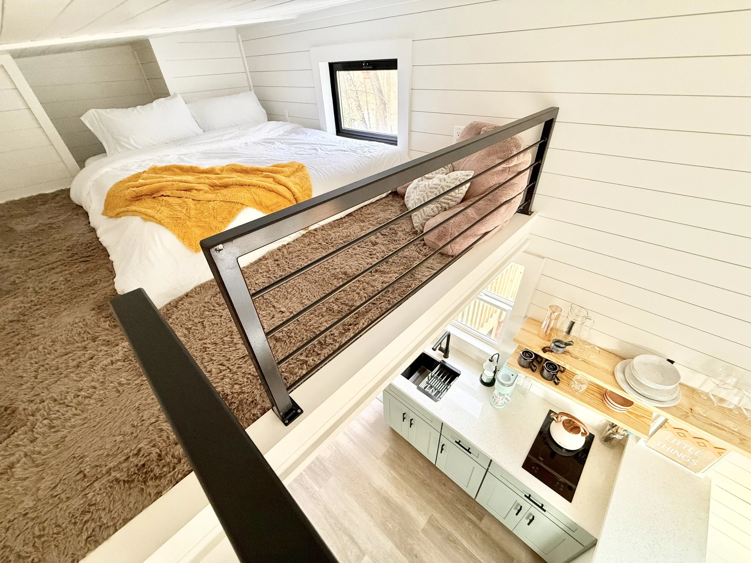A cozy upstairs bedroom with a bed covered in white bedding and an orange throw blanket, a small window, and a railing overlooking a kitchen below with light green cabinets, white countertop, and wooden shelves holding dishes and glassware.