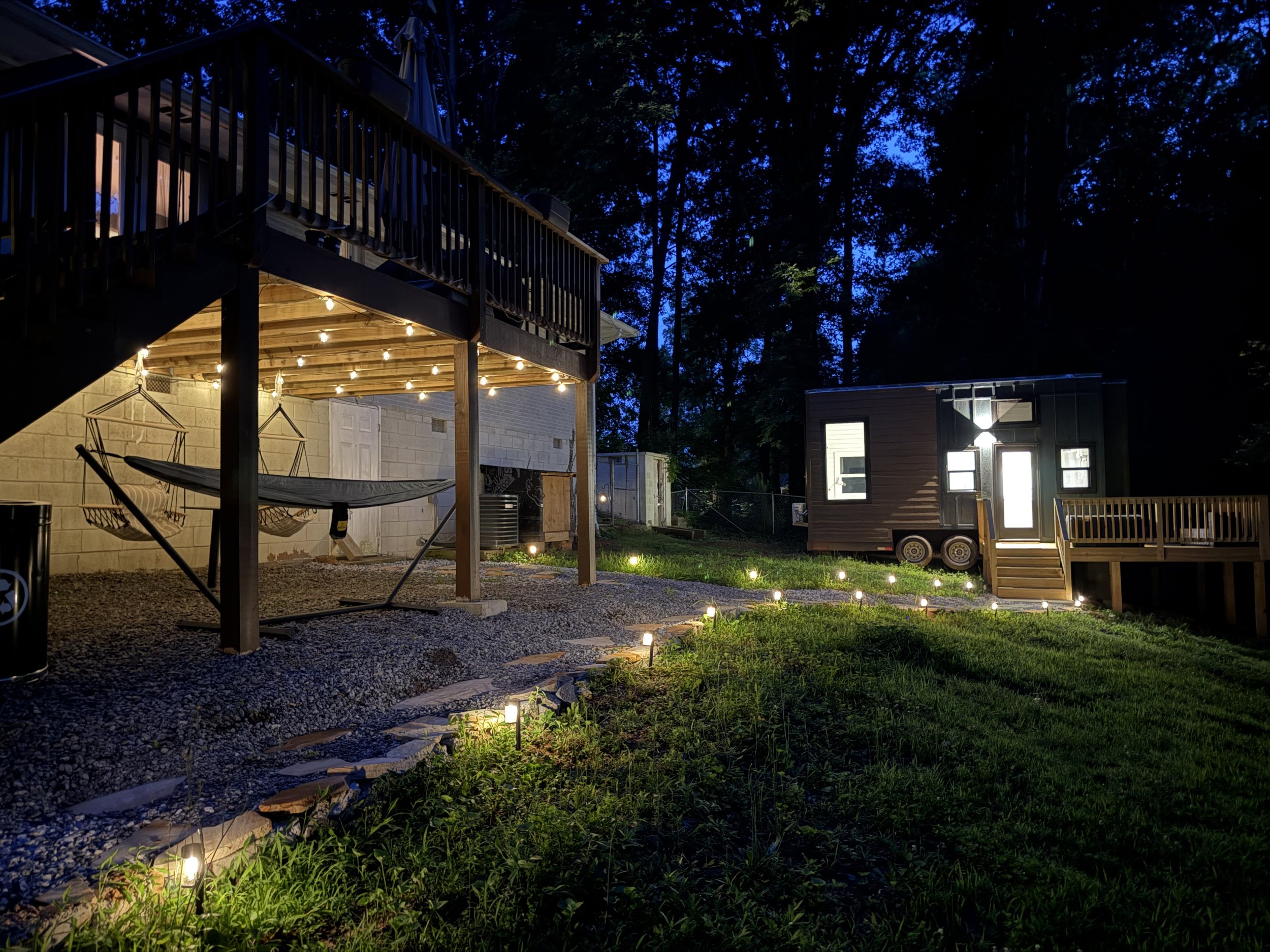 A backyard at night with outdoor lighting, a wooden deck with string lights, a hammock, a small building with lit windows, and trees in the background.