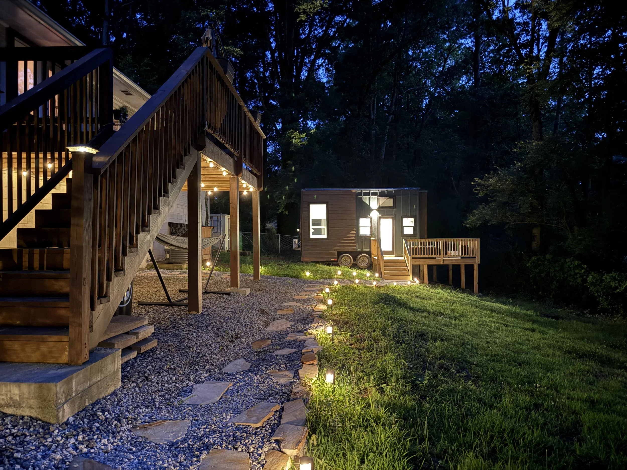 Nighttime backyard scene with a wooden staircase, string lights, a small modern shed, and illuminated path lights on a grassy yard surrounded by trees.