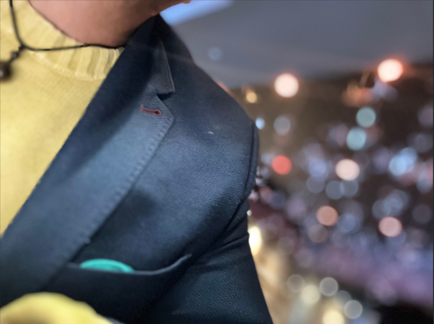 Close-up of a person wearing a yellow shirt and a dark blazer, with blurred bokeh lights in the background.