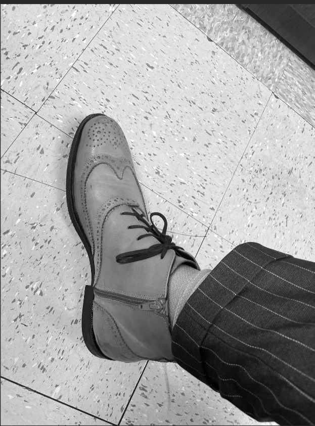 A black-and-white photo of a person's leg and foot, wearing a dress shoe and pinstripe pants, with the foot resting on a tiled floor.