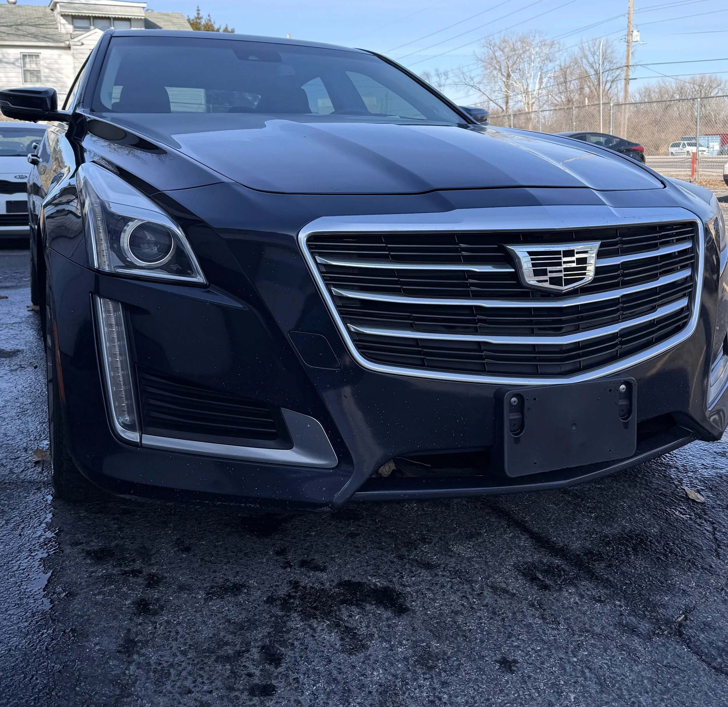 Front view of a black Cadillac sedan parked outdoors on a wet surface, with other vehicles, a building, and a chain-link fence in the background.