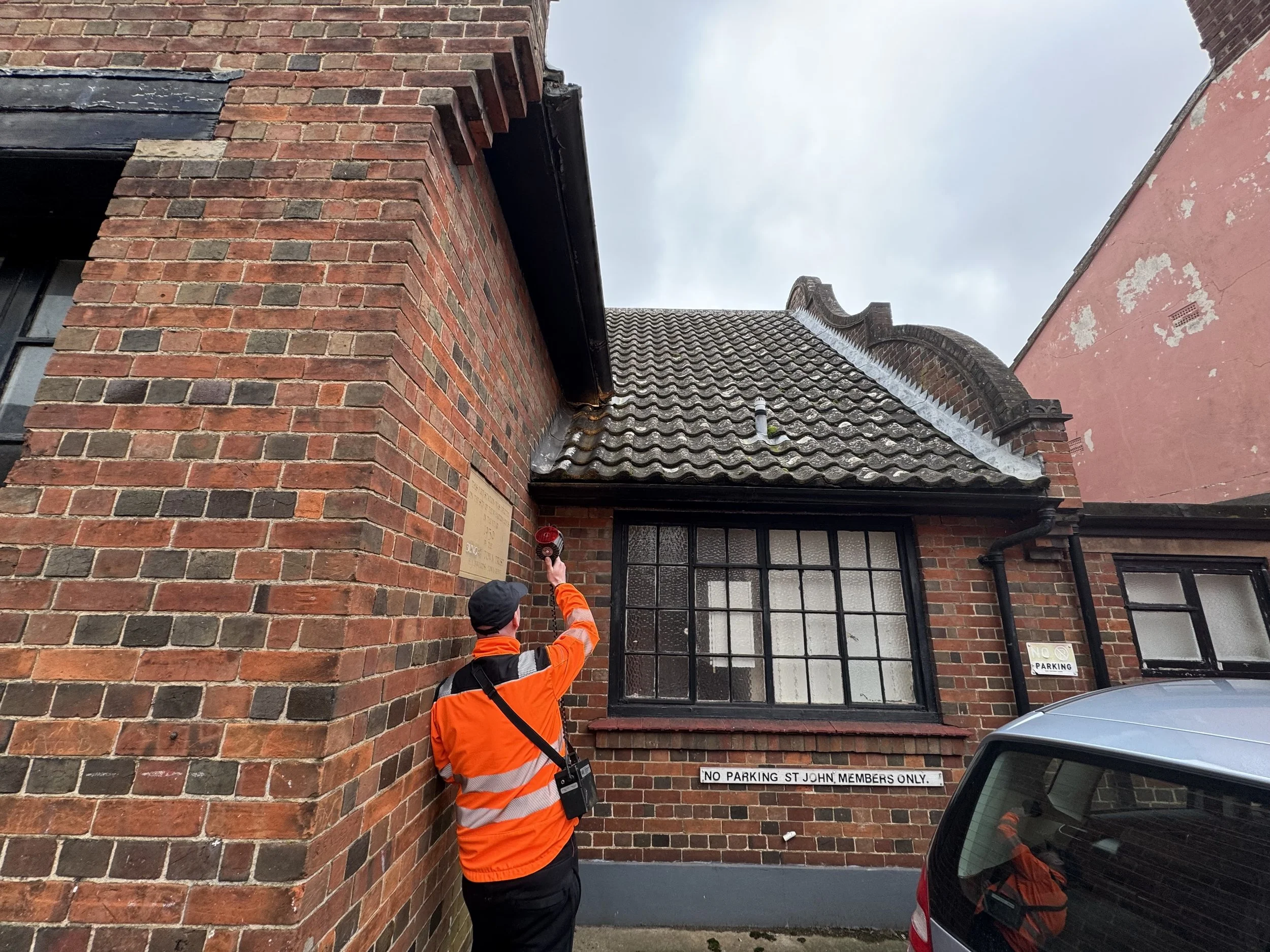 A worker in an orange safety vest is ringing a bell on a brick building. The building has a sign that reads 'NO PARKING ST JOHN MEMBERS ONLY.' There is a dark-colored car parked in front of the building and a cloudy sky above.