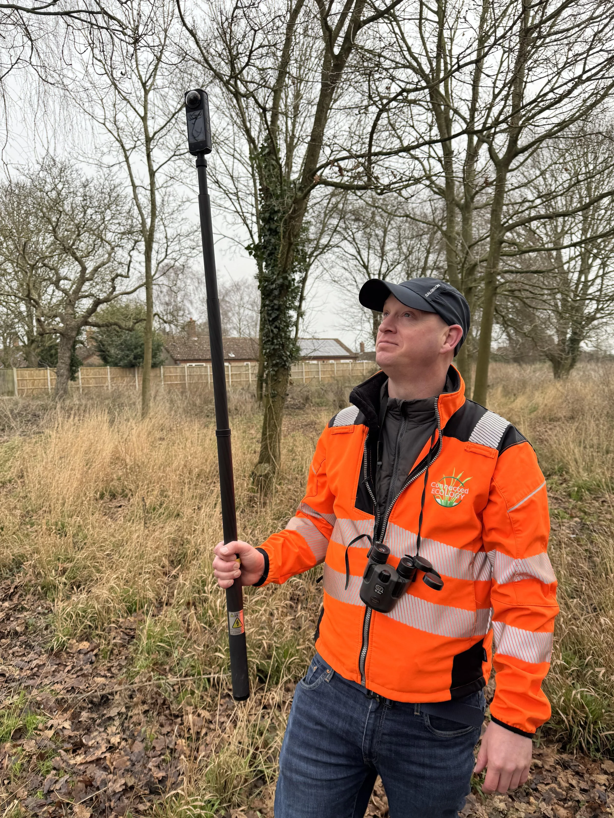 A man wearing an orange high-visibility jacket with binoculars around his neck, holding a long monopod with a camera or sensor, standing outdoors in a wooded area on a cloudy day.