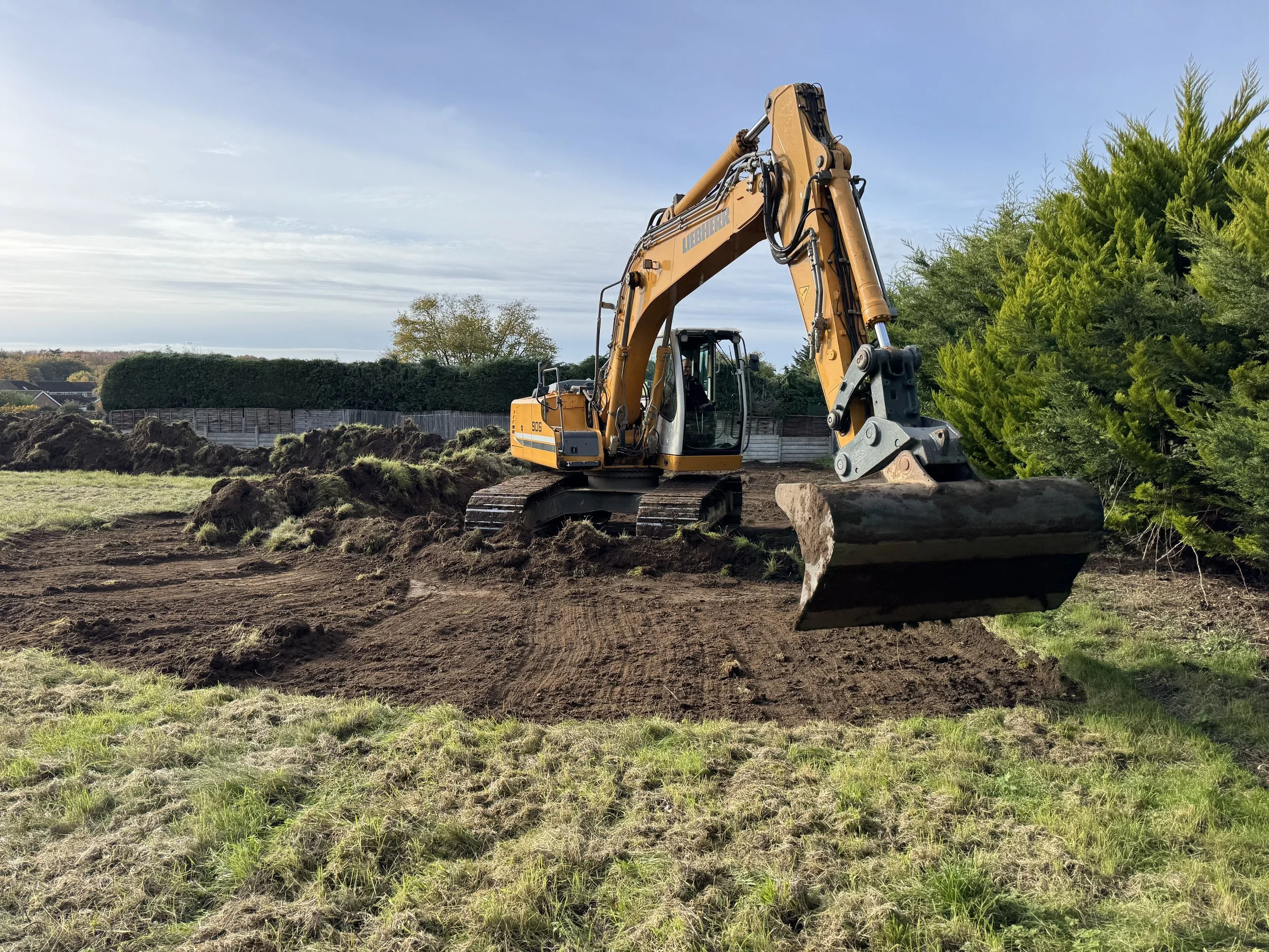 A yellow excavator on a grassy field digging up soil with a bucket, with trees and a fence in the background.