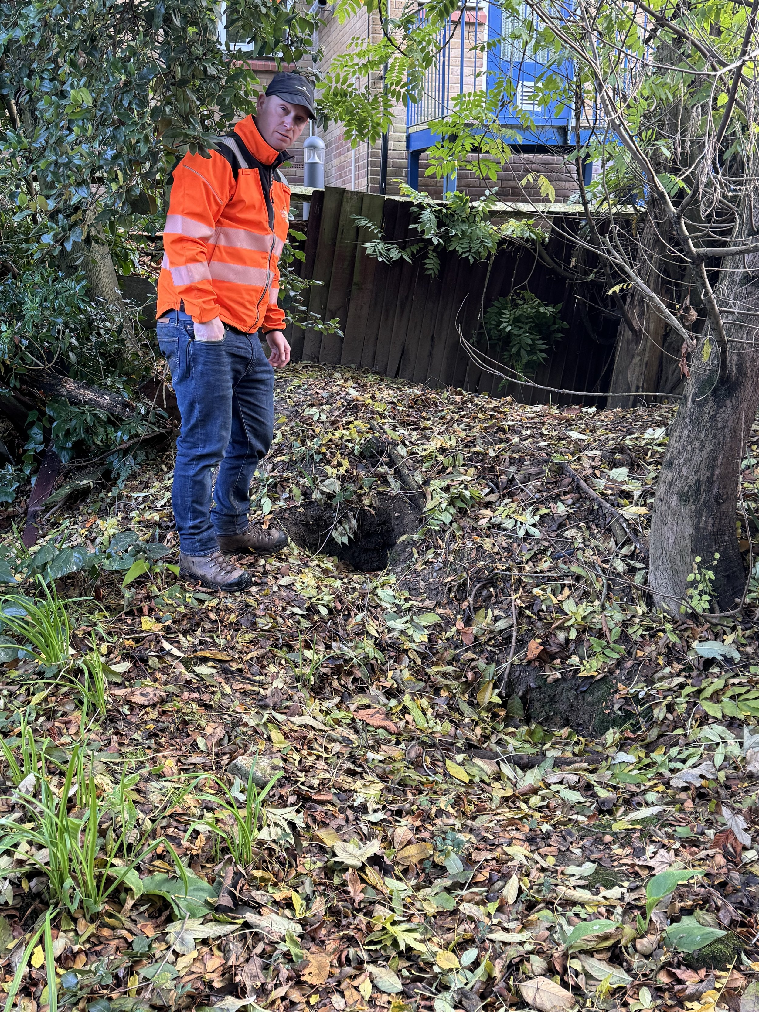 A person in a bright orange safety jacket and jeans standing next to a small hole in the ground in a wooded backyard.