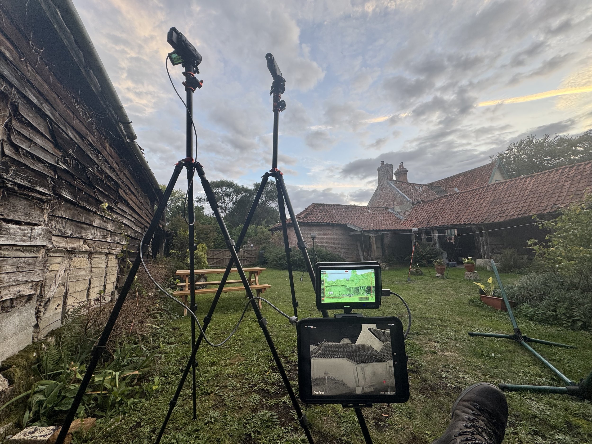 Recording equipment set up in a backyard with a rustic wooden shed, in front of a house with red-tiled roof, during sunset with cloudy sky.