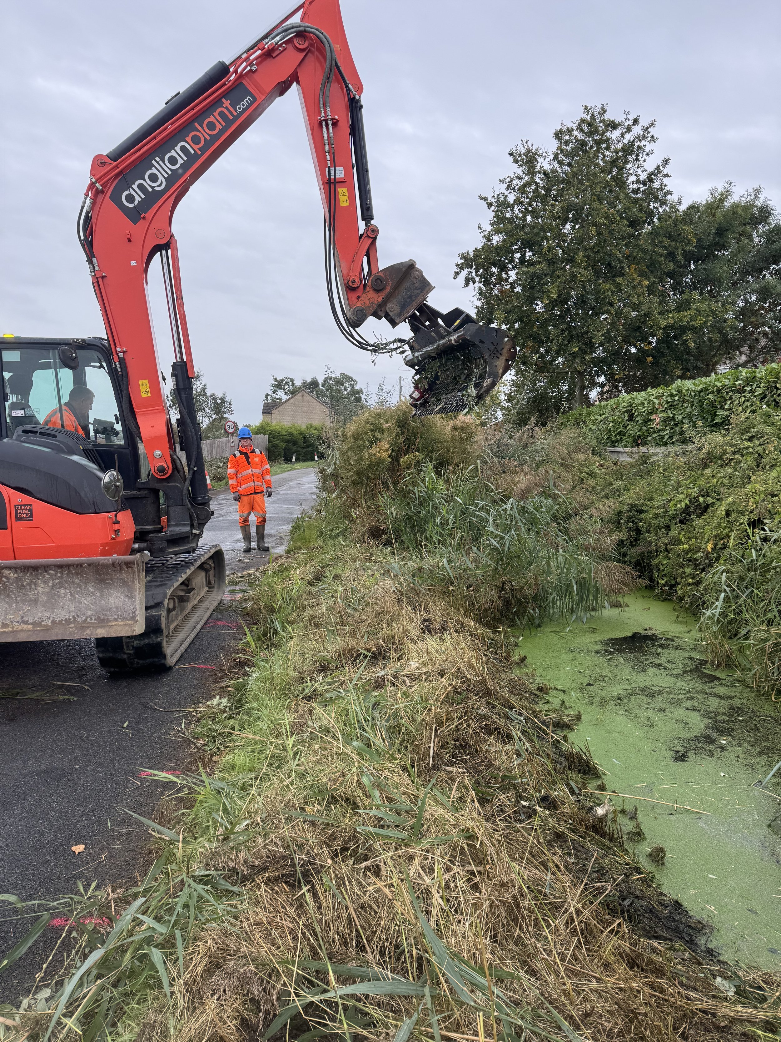 A large red excavator with the text angliaplant.com on its arm is removing overgrown bushes and plants from a roadside ditch. Two workers in orange safety vests and helmets are nearby, working under an overcast sky.