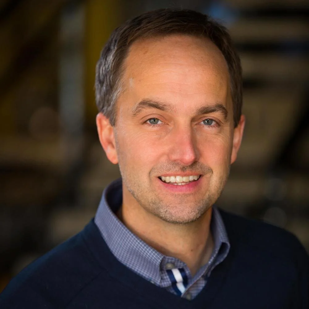 A smiling man with short brown hair, blue eyes, and a light beard, wearing a collared shirt and a dark sweater, standing in a blurred indoor background.