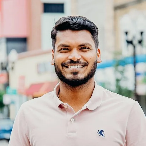 A smiling man with dark hair and a beard, wearing a light pink polo shirt with a small blue lion emblem, standing outdoors in an urban area.