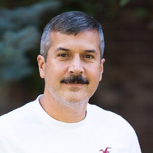 A man with short gray hair and a mustache wearing a white shirt, outdoors with blurred greenery in the background.