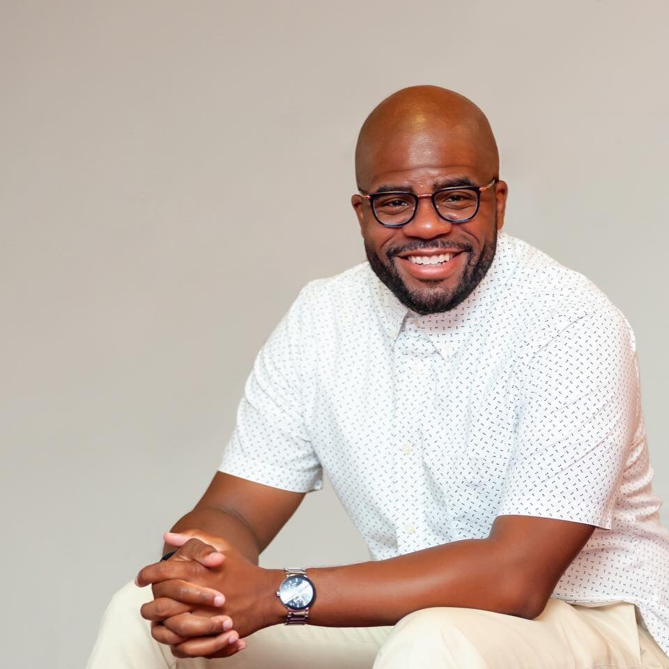 A smiling man with glasses and a beard, wearing a white dotted shirt and a watch, sitting and clasping his hands, against a plain light-colored background.