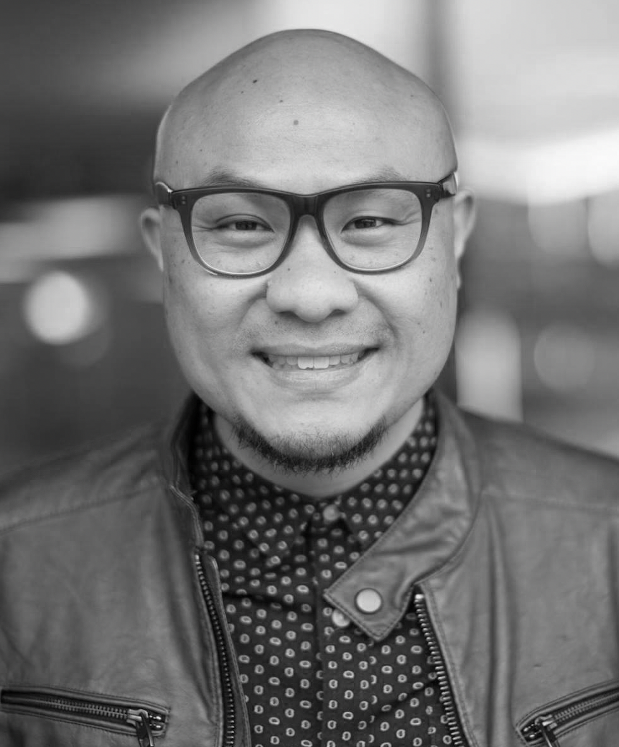 Black and white portrait of a smiling man wearing glasses, a patterned shirt, and a leather jacket.