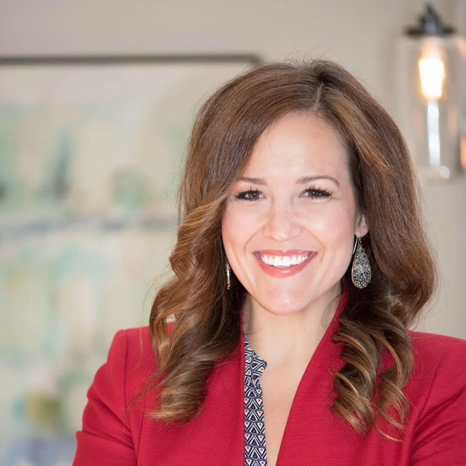A woman with long brunette hair smiling, wearing a red blazer and patterned blouse, in a well-lit indoor setting.