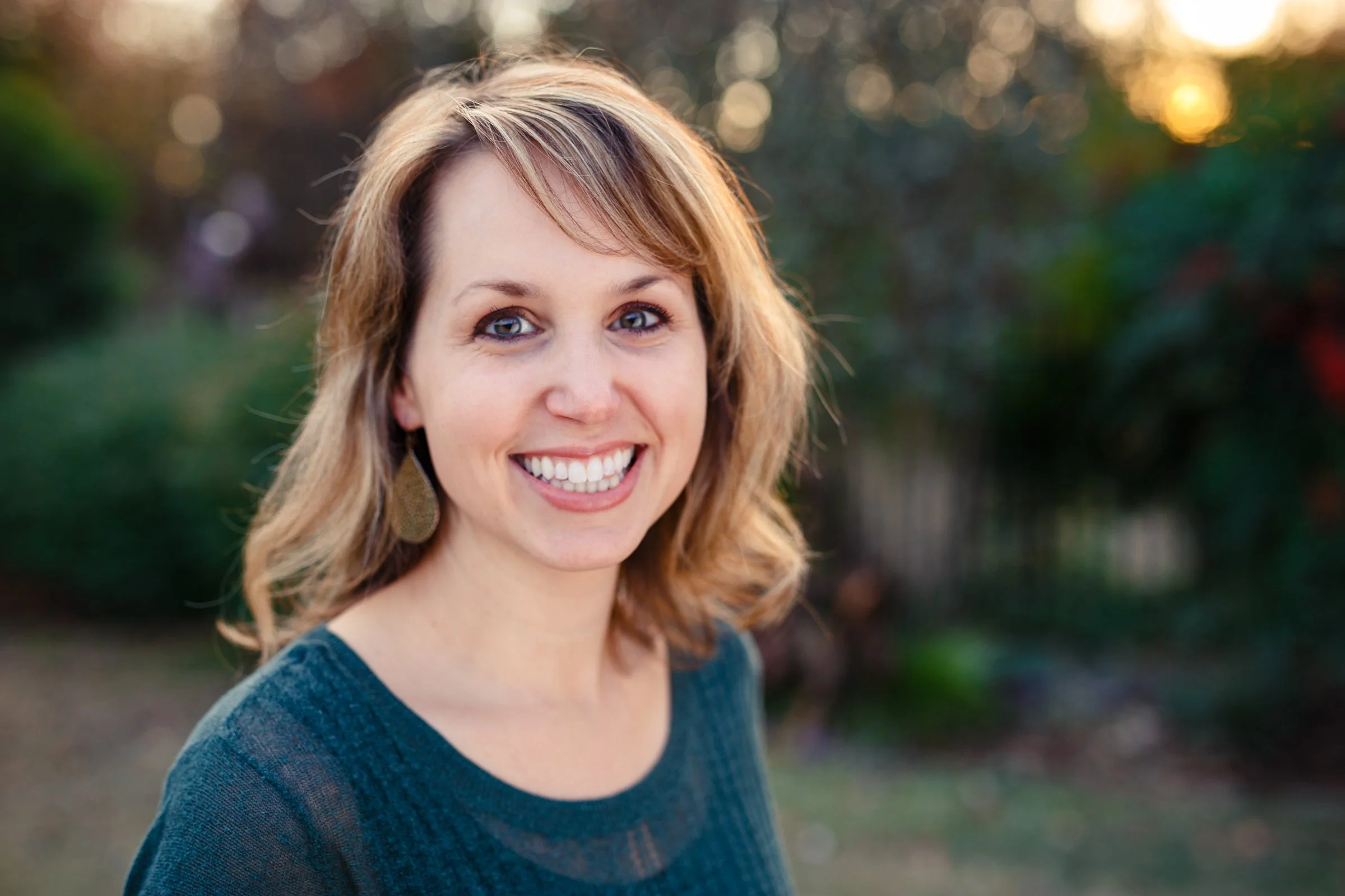 A woman with shoulder-length light brown hair, smiling with white teeth, wearing a dark blue top outdoors during sunset with blurred greenery in the background.