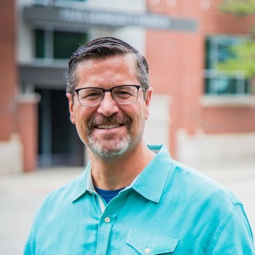 A middle-aged man with short gray hair, glasses, and a beard, smiling outdoors in front of a brick building.