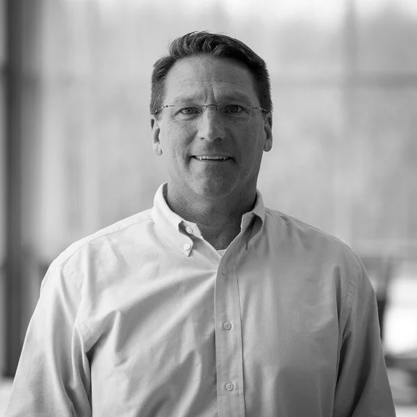 Black and white photo of a man with glasses, wearing a button-up shirt, smiling, in an indoor setting.