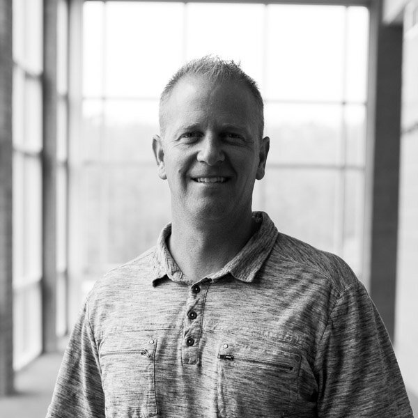 A smiling man standing indoors in front of large windows, wearing a casual button-up shirt.