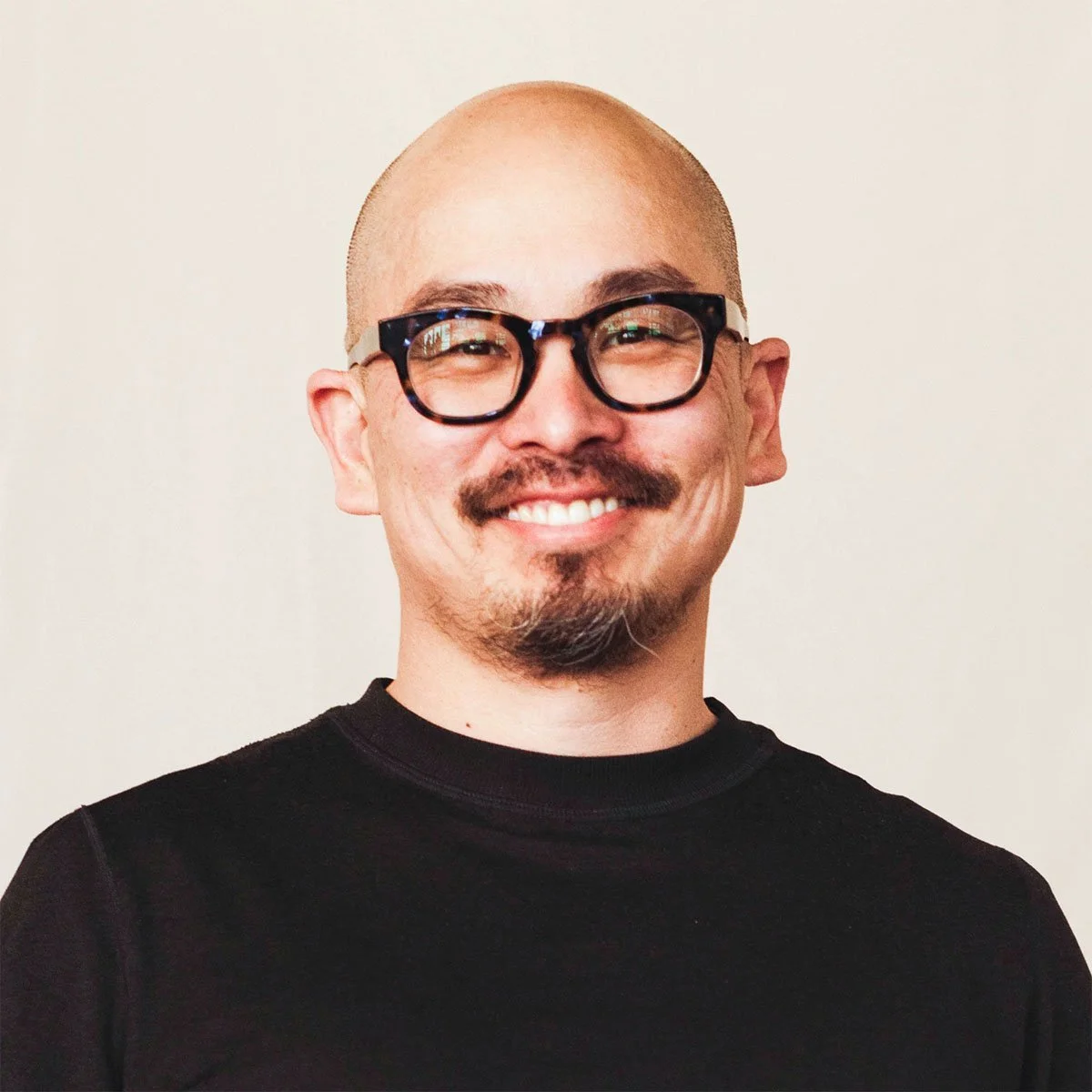 A smiling man with glasses, a mustache, and a goatee, wearing a black shirt, standing against a plain light-colored background.