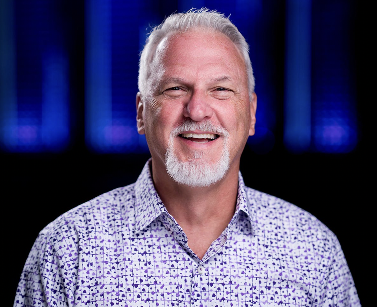 A smiling older man with white hair and beard wearing a purple patterned shirt, posing in front of a blue-lit background.