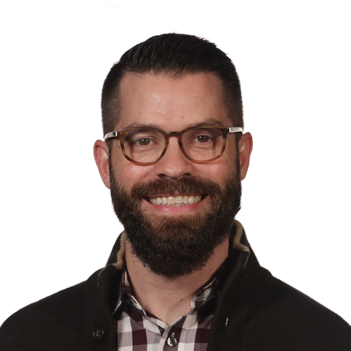 A smiling man with dark hair, glasses, a beard, and a mustache wearing a dark jacket over a plaid shirt, against a white background.