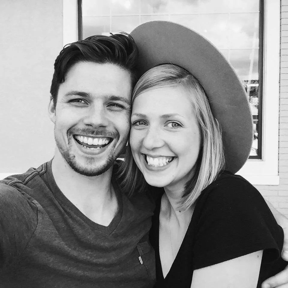 A smiling man and woman taking a selfie, with the woman wearing a wide-brimmed hat, in front of a building with a window.